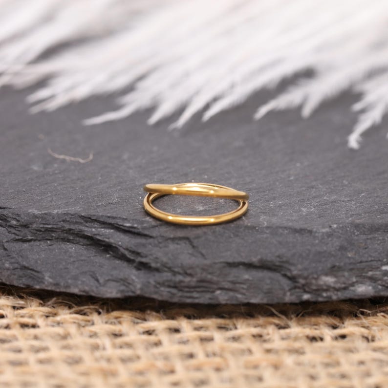 May include: A gold-colored ring with a double-band design. The ring is made of a shiny, reflective metal and sits on a dark gray surface. The ring's bands are smooth and rounded, creating a minimalist and elegant look.
