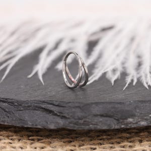 May include: A silver-colored ring with a double-band design. The bands intersect, creating a unique, modern look. The ring is set against a dark gray surface with white, feathery accents in the background, and a burlap texture.