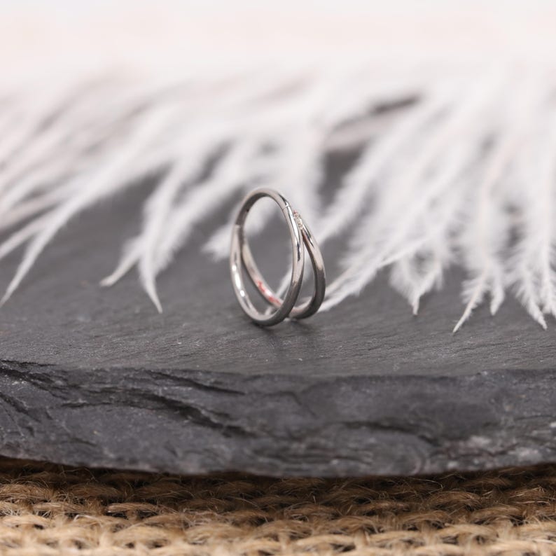 May include: A silver-colored ring with a double-band design. The bands intersect, creating a unique, modern look. The ring is set against a dark gray surface with white, feathery accents in the background, and a burlap texture.
