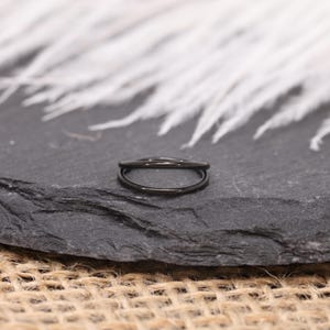 May include: A black double-band ring, likely made of metal, sits on a dark gray stone surface. The ring's design features two parallel bands, creating a sleek, modern look. The background includes white feathers and a textured surface.