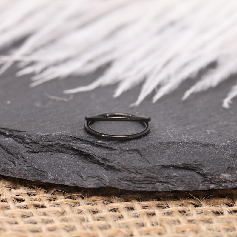 May include: A black double-band ring, likely made of metal, sits on a dark gray stone surface. The ring's design features two parallel bands, creating a sleek, modern look. The background includes white feathers and a textured surface.