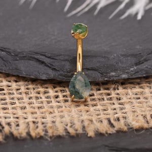 Puede incluir: Un piercing para el ombligo de color dorado con dos piedras preciosas verdes. La piedra superior es redonda y la inferior tiene forma de lágrima. La joya se exhibe sobre una superficie gris oscuro con un fondo de arpillera.