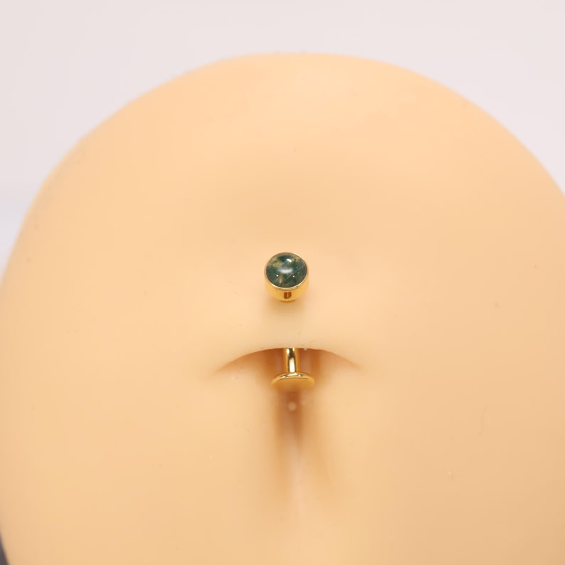 May include: A gold-coloured belly button ring featuring a round, green stone. The jewellery is shown on a mannequin. The belly button ring has a post and a small, flat disc on the bottom.