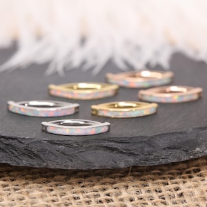 May include: Six rings with a rectangular opal inlay are displayed on a dark surface. The rings have different metal finishes: silver, gold, and rose gold. The opals have a white base with iridescent flashes of color.