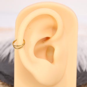 May include: A gold-colored double hoop earring is shown on a tan ear model. The earring is a small, circular design with two parallel hoops. The ear model is set against a blurred background.
