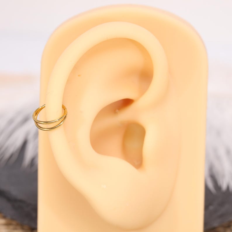 May include: A gold-colored double hoop earring is shown on a tan ear model. The earring is a small, circular design with two parallel hoops. The ear model is set against a blurred background.