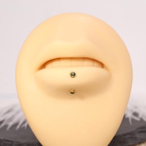 May include: A close-up of a tan-colored mouth model with two pieces of jewelry. The top piece is a small, round, gold-colored stud with a green gem. The bottom piece is a similar gold-colored stud with a green gem.