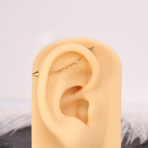 Pode incluir: Um piercing industrial de tom dourado com duas extremidades cónicas pontiagudas e uma delicada corrente de elos suspensa entre as duas barras, exibido num modelo de orelha bege para mostrar a colocação através da cartilagem superior.