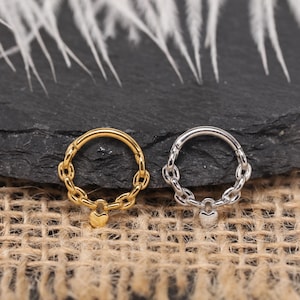 May include: Two hinged nose rings. One is gold-toned, the other is silver-toned. Both feature a chain design around the hoop and a small heart charm. The jewelry is displayed on a textured surface.