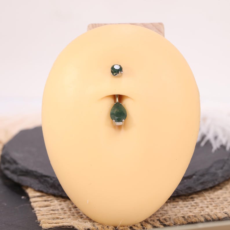May include: A belly button ring with a teardrop-shaped green gemstone pendant. The jewelry is silver-toned and features a small, round green gemstone on the top. The jewelry is displayed on a tan-colored, oval-shaped form.