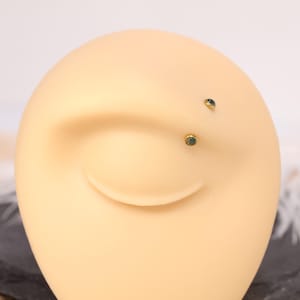 May include: Close-up of a tan-colored ear model with two small, gold-colored stud earrings. Each earring features a small, round, green gemstone. The earrings are positioned on the earlobe and upper ear.