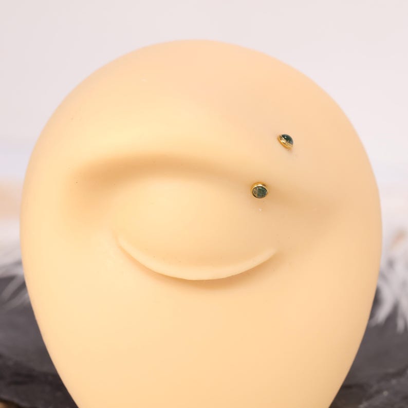 May include: Close-up of a tan-colored ear model with two small, gold-colored stud earrings. Each earring features a small, round, green gemstone. The earrings are positioned on the earlobe and upper ear.