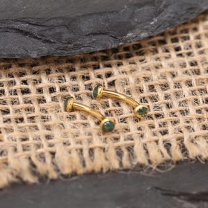 May include: Two curved, gold-colored eyebrow rings with small, round, green gemstones at each end. The jewelry is displayed on a textured, tan fabric background, with a dark gray surface in the upper portion of the image.