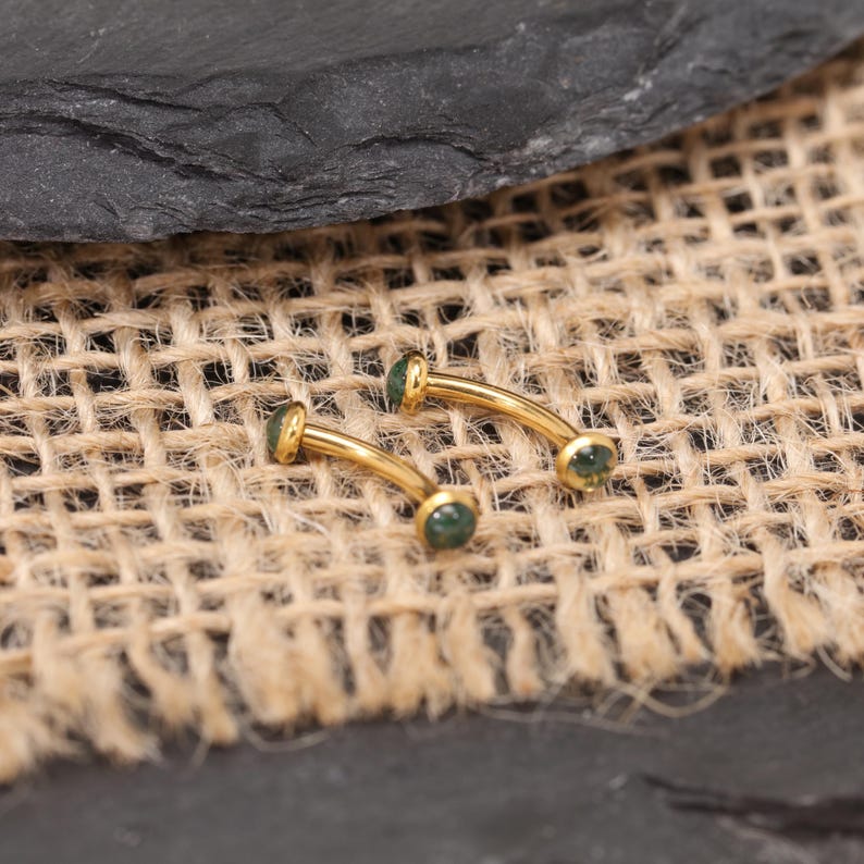 May include: Two curved, gold-colored eyebrow rings with small, round, green gemstones at each end. The jewelry is displayed on a textured, tan fabric background, with a dark gray surface in the upper portion of the image.