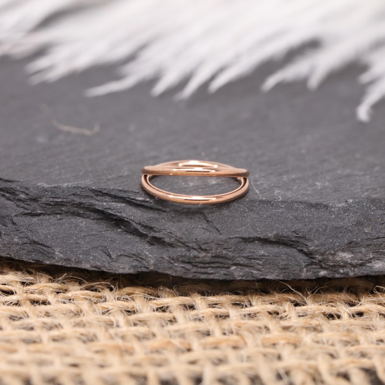May include: A rose gold double hoop ring. The ring is set on a dark gray stone surface with a blurred white and gray background. The ring's design features two parallel, curved bands.