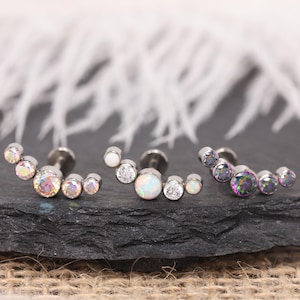 Puede incluir: Tres pares de pendientes con un diseño curvo. Cada pendiente presenta una serie de pequeñas piedras preciosas redondas en colores iridiscentes, blancos y transparentes. Los pendientes están engastados en una base de metal plateado, expuestos sobre una superficie oscura y texturizada.