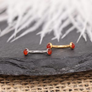 May include: Two curved barbells with red gemstones. One barbell is silver, and the other is gold. The gemstones are oval-shaped and set in a bezel setting. The jewellery is displayed on a dark grey surface.