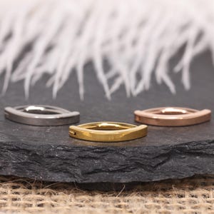 Puede incluir: Tres anillos de metal curvados en plata, oro y oro rosa. Los anillos se muestran sobre una superficie gris oscuro con un fondo blanco borroso. Los anillos tienen un diseño minimalista.