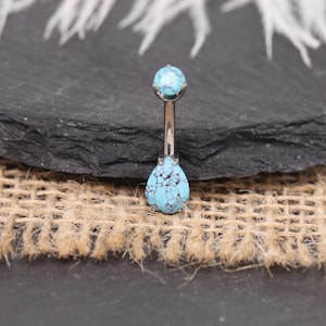 Op de afbeelding: Een zilveren navelpiercing met turkooizen stenen. De bovenste steen is rond en de onderste steen is druppelvormig. Het turquoise heeft een blauw en zwart gespikkeld patroon. De ring wordt gepresenteerd op een donkergrijze stenen ondergrond.
