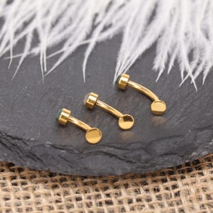 May include: Three gold-coloured curved eyebrow piercing jewellery pieces with small, round, green and clear gemstones. The jewellery is displayed on a dark grey surface with white feathers in the background. The jewellery has a curved barbell design.