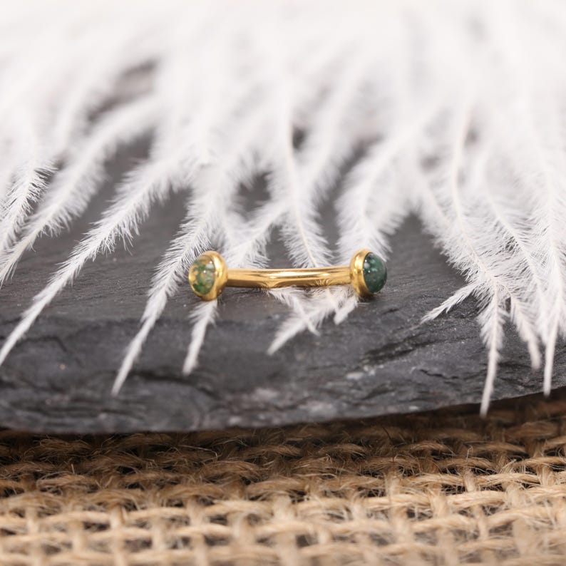 May include: A gold-colored barbell earring with green gemstones at each end. The earring is lying on a dark gray surface with white feathers in the background. The earring is a small, delicate piece of jewelry.