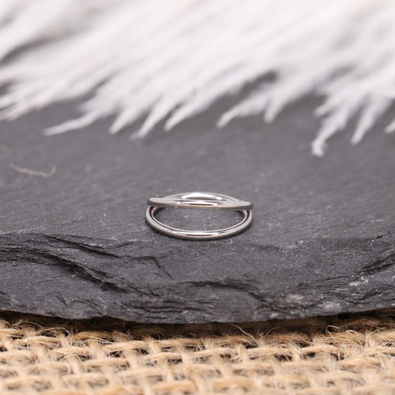 May include: A silver-colored double-band ring. The ring has a smooth, polished finish and a minimalist design. The ring is set against a dark gray, textured surface.