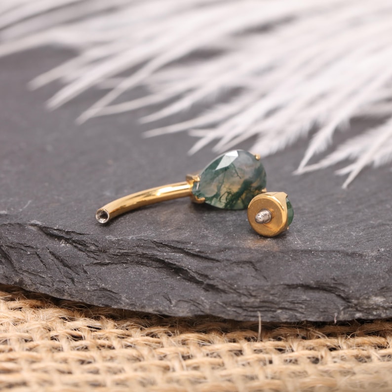 May include: A gold-colored, curved barbell body piercing with a teardrop-shaped, green and white gemstone. A matching gold-colored end piece with a small, clear gem is also present. The jewelry is set against a dark, textured background.