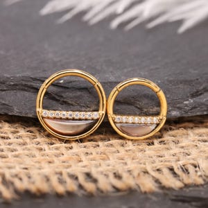 Op de afbeelding: Twee goudkleurige oorringen. Elke oorring heeft een halve cirkel van iriserend materiaal en een rij kleine, heldere edelstenen. De oorbellen zijn geplaatst tegen een donkere, getextureerde achtergrond.