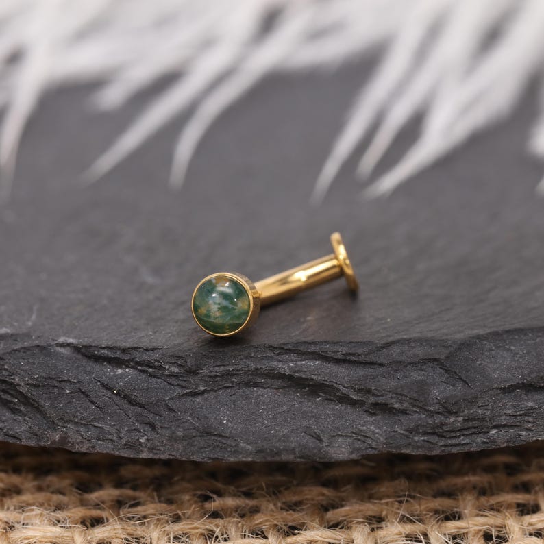May include: A gold-coloured, flat-back earring featuring a round, green stone. The earring has a straight post and a small, flat disc at the back. The stone has a mottled appearance with shades of green.