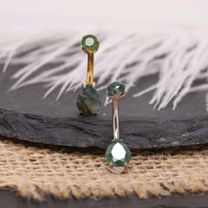 May include: Two belly button rings with green gemstones. One ring has a gold-colored curved bar with a teardrop-shaped gemstone and a small round gemstone. The other ring has a silver-colored bar with a faceted gemstone.