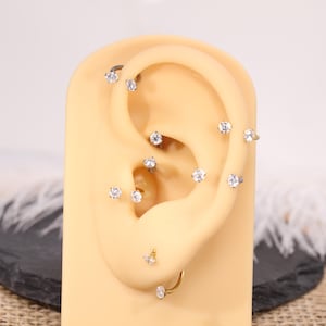 Könnte beinhalten: Darstellung verschiedener Ohrpiercings, darunter Stecker und Creolen, die mit klaren, funkelnden Edelsteinen besetzt sind. Die Piercings sind auf einem beigefarbenen Ohrmodell angebracht, das die Platzierung und das Design des Schmucks zeigt.