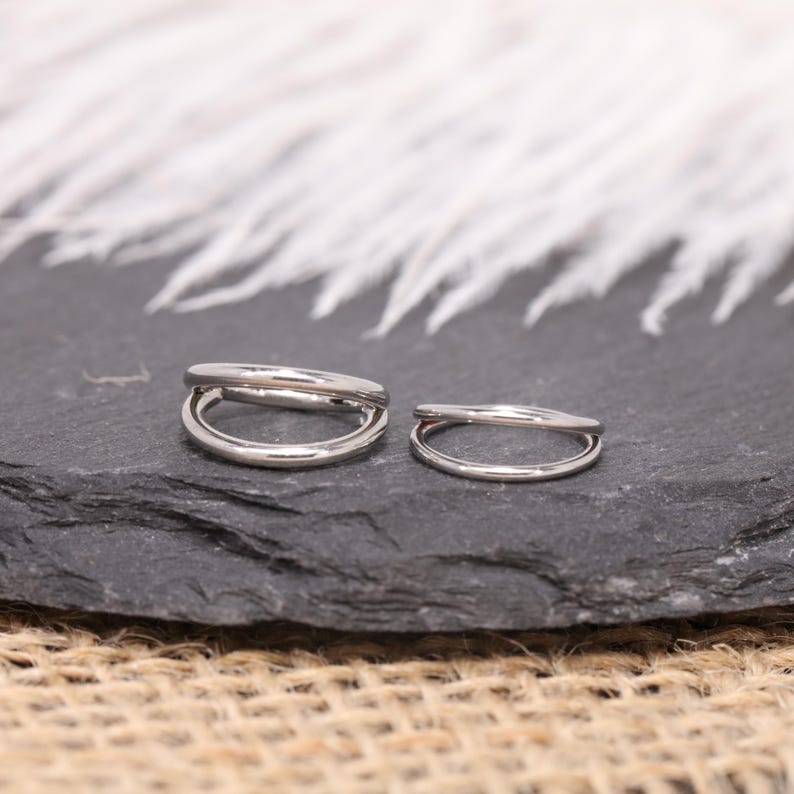 May include: Two silver-colored, double-banded ear cuffs. The ear cuffs are displayed on a dark gray, textured surface, with a blurred white background. The ear cuffs are small and minimalist in design.