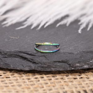 May include: A small, iridescent ring with a double band design. The ring displays a spectrum of colors, including green, blue, and purple. The ring is set against a dark, textured background, with a blurred, white feathery element in the background.
