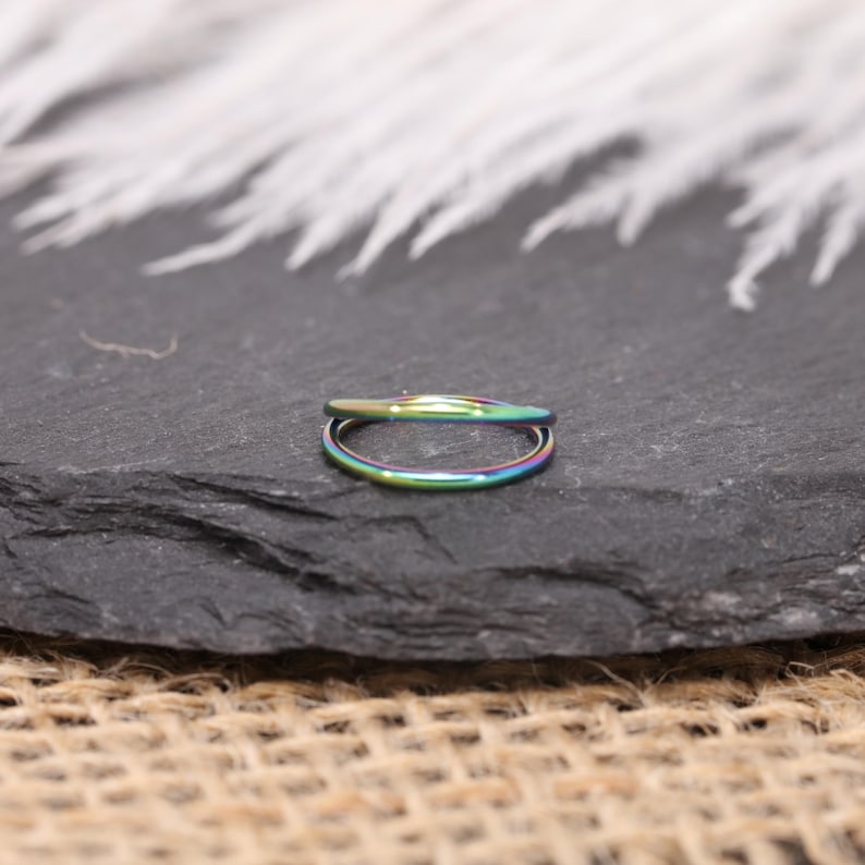 May include: A small, iridescent ring with a double band design. The ring displays a spectrum of colors, including green, blue, and purple. The ring is set against a dark, textured background, with a blurred, white feathery element in the background.