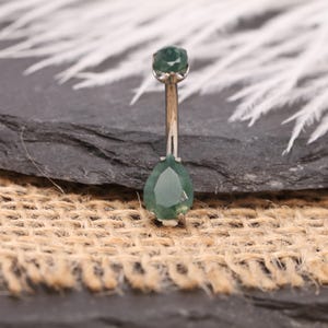 May include: A belly button ring with a silver-colored bar and two green, faceted gemstones. The top gem is round, and the bottom gem is teardrop-shaped. The jewelry is displayed on a dark gray surface with a burlap texture.