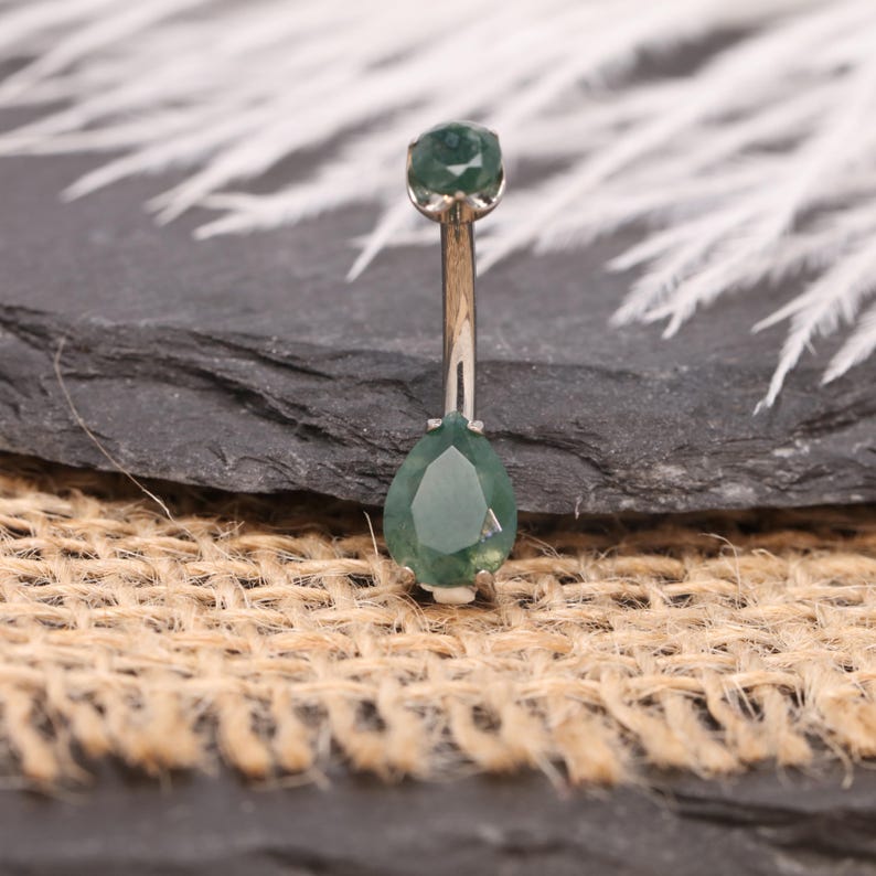 May include: A belly button ring with a silver-colored bar and two green, faceted gemstones. The top gem is round, and the bottom gem is teardrop-shaped. The jewelry is displayed on a dark gray surface with a burlap texture.