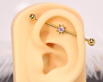 Puede incluir: Pendiente industrial barbell dorado con un diseño de estrella y una piedra central morada. El pendiente se muestra en un modelo de oreja bronceada, mostrando el estilo y la colocación de la joya.