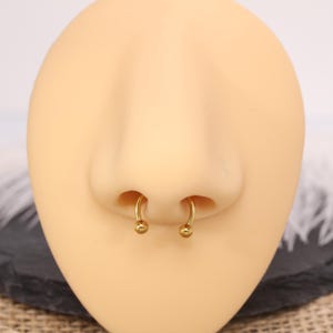 May include: A close-up of a nose with a gold-colored septum ring. The ring is a horseshoe shape with a small ball on each end. The skin tone is light.