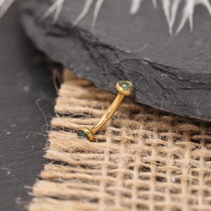 May include: A curved, gold-colored eyebrow ring with two small, round, green gemstones. The jewelry is displayed on a textured, neutral-colored surface with a dark gray stone in the background.
