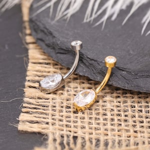 Piercing de umbigo oval com zircônia cúbica branca 14G, em titânio, ouro e prata, com base plana. Design delicado e minimalista.