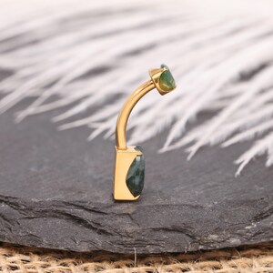 May include: A gold-colored belly button ring with a curved bar. The ring features a rectangular green gemstone at the bottom and a smaller, rounded green gemstone at the top. The jewelry is displayed on a dark gray surface.