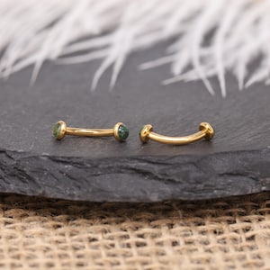 May include: Two curved, gold-colored eyebrow piercings with small, round, green gemstones at each end. The jewelry is displayed on a dark gray, textured surface, with a blurred white background.
