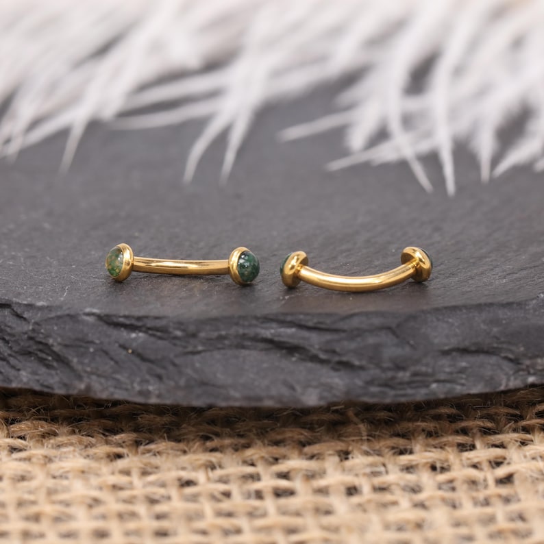 May include: Two curved, gold-colored eyebrow piercings with small, round, green gemstones at each end. The jewelry is displayed on a dark gray, textured surface, with a blurred white background.