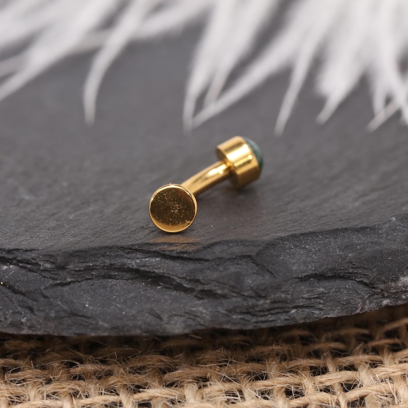 May include: A gold-coloured, curved barbell earring. It has a small, flat disc on one end and a small, round, green stone on the other. The earring is on a dark, textured surface.