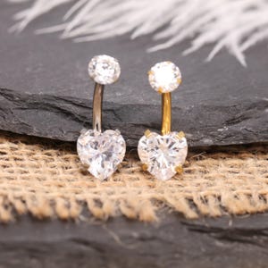 May include: Two belly button rings with heart-shaped and round clear gemstones. One ring has a silver-colored curved bar and the other has a gold-colored bar. The jewelry is displayed on a dark gray surface with a burlap texture.