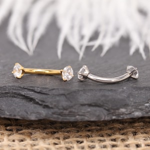 May include: Two curved eyebrow rings with clear, round gemstones. One ring is gold-colored, and the other is silver-colored. The jewelry is displayed on a dark gray surface with a blurred white background.