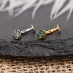 May include: Two flat-back earrings. One is silver-toned, the other gold-toned. Each has a small, round, green and white stone set in a bezel. The earrings are displayed on a dark, textured surface.