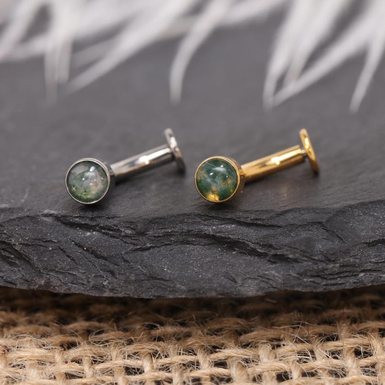 May include: Two flat-back earrings. One is silver-toned, the other gold-toned. Each has a small, round, green and white stone set in a bezel. The earrings are displayed on a dark, textured surface.