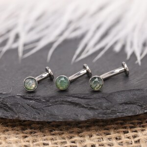 May include: Three silver labret studs with green gemstones. The studs are displayed on a dark, textured surface. The background is blurred and white. The gemstones have a green hue.