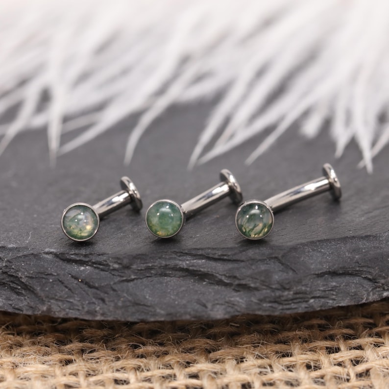 May include: Three silver labret studs with green gemstones. The studs are displayed on a dark, textured surface. The background is blurred and white. The gemstones have a green hue.